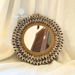 Round decorative mirror with a woven natural fiber frame and a border of small seashells, displayed against draped white fabric. Tropea Artistry Bali watermark is visible in the upper left.