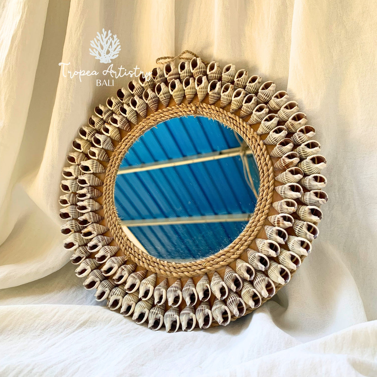 Round decorative wall mirror with a woven natural fiber frame and a border of small seashell-like accents, shown draped against cream fabric with blue reflected in the mirror.