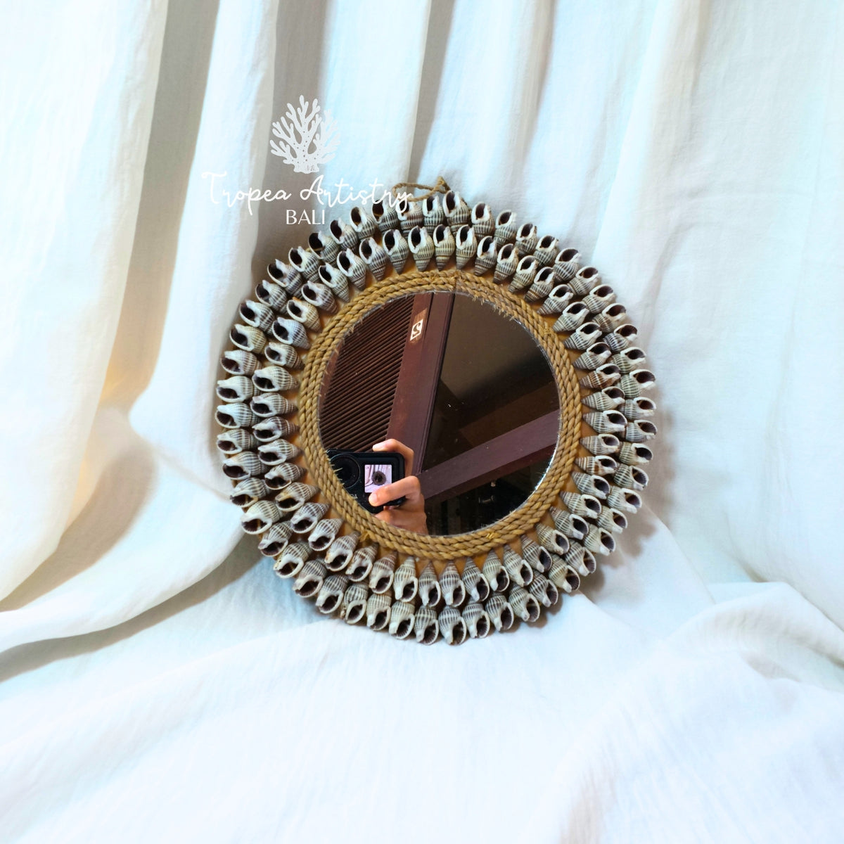 A round decorative mirror with a wide, textured frame made of many small silver-toned circular pieces around a natural wood center, resting against draped white fabric. The mirror reflects a person taking a photo, and a watermark for 'Toppea Artistry' is visible on the upper left.