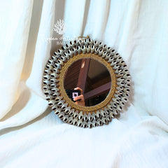 A round decorative mirror with a wide, textured frame made of many small silver-toned circular pieces around a natural wood center, resting against draped white fabric. The mirror reflects a person taking a photo, and a watermark for 'Toppea Artistry' is visible on the upper left.
