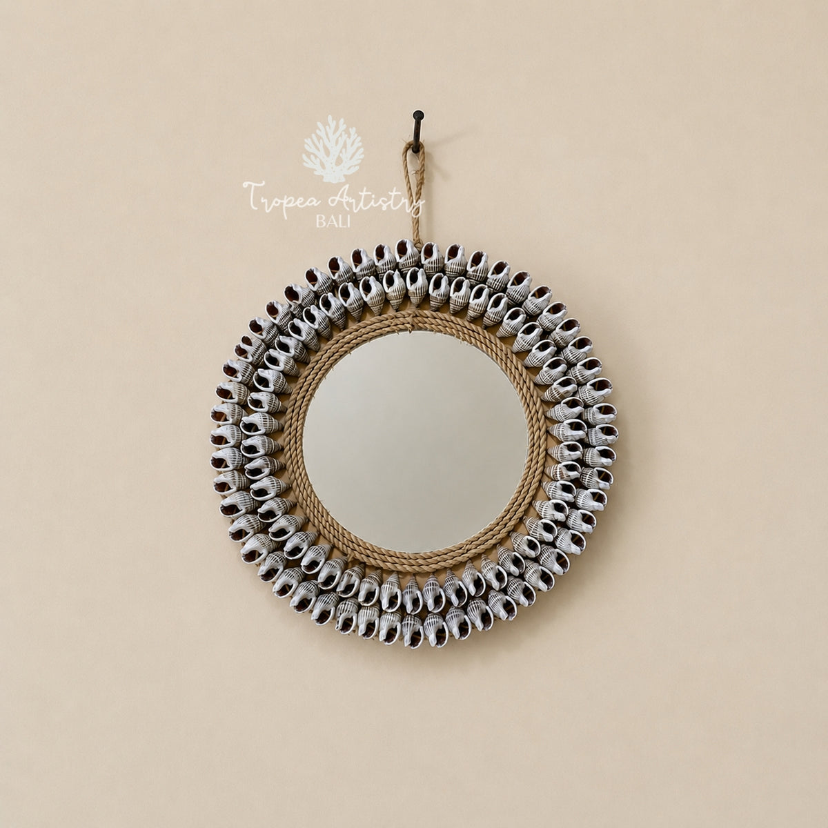A round wall mirror with a decorative border made of small silver bells and natural jute rope, hanging on a beige wall with a visible Tropê Artistry Balls watermark.