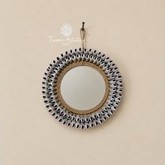 A round wall mirror with a decorative border made of small silver bells and natural jute rope, hanging on a beige wall with a visible Tropê Artistry Balls watermark.