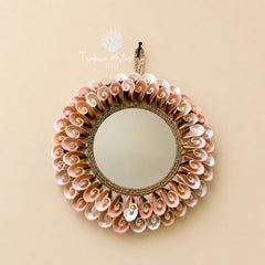 A round wall mirror with a decorative border made of layered seashells in soft pink, beige, and white tones, wrapped with natural rope. The mirror is shown hanging against a solid beige wall, with a 'Tropica Artistry Bali' logo watermark above it.