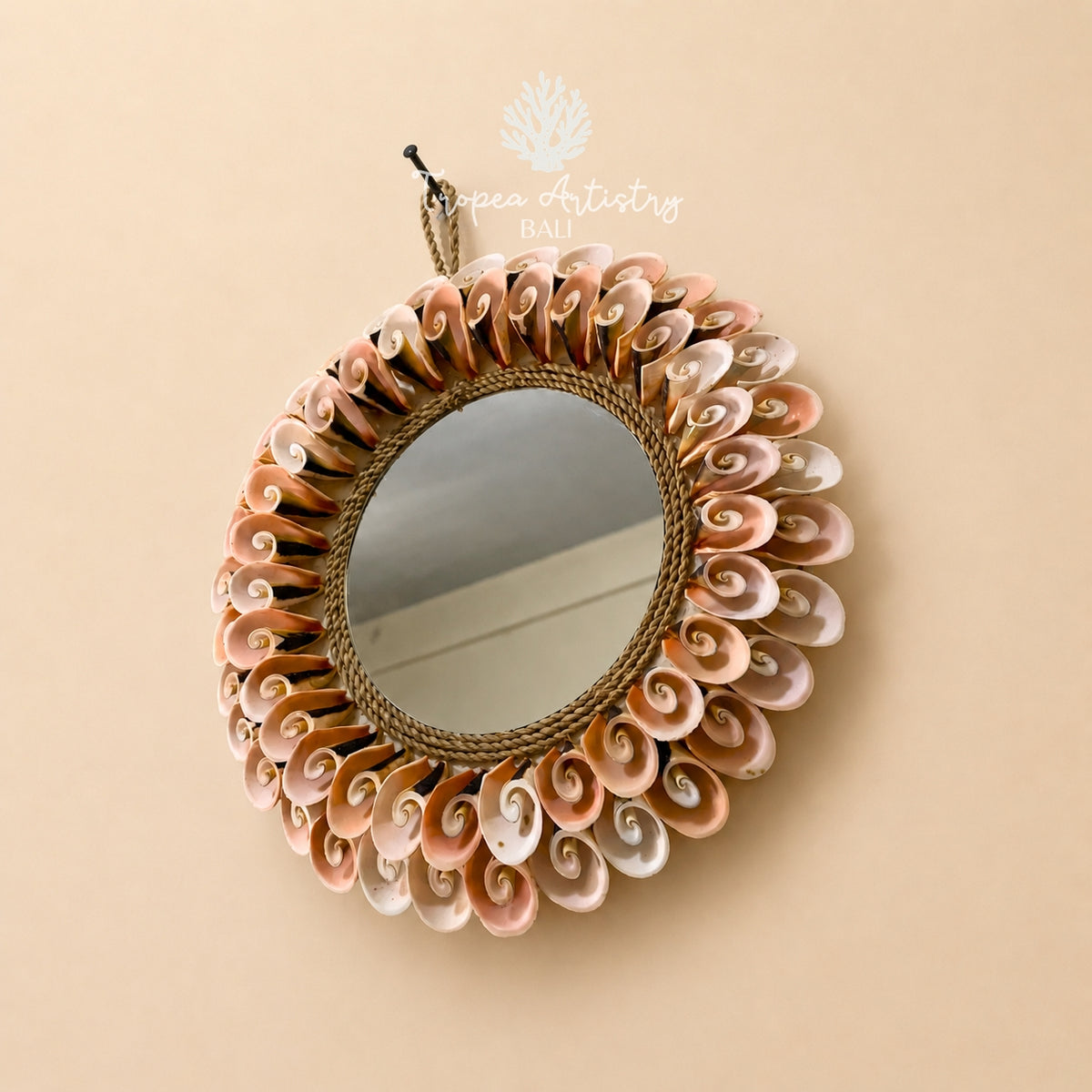 An oval wall mirror with a decorative sunburst frame made of layered seashells in soft pink, beige, and white tones, surrounded by a braided rope border. The mirror is hanging against a solid beige wall, with a 'pea Artistry Bali' watermark at the top.