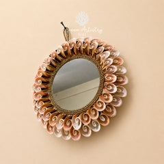 An oval wall mirror with a decorative sunburst frame made of layered seashells in soft pink, beige, and white tones, surrounded by a braided rope border. The mirror is hanging against a solid beige wall, with a 'pea Artistry Bali' watermark at the top.