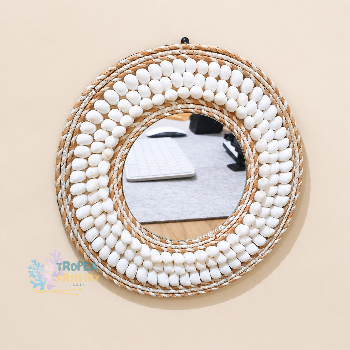 A round decorative wall mirror with a thick rope border and rows of white pebble-like accents, mounted on a beige wall. The reflection shows a desk setup with a keyboard and mouse.