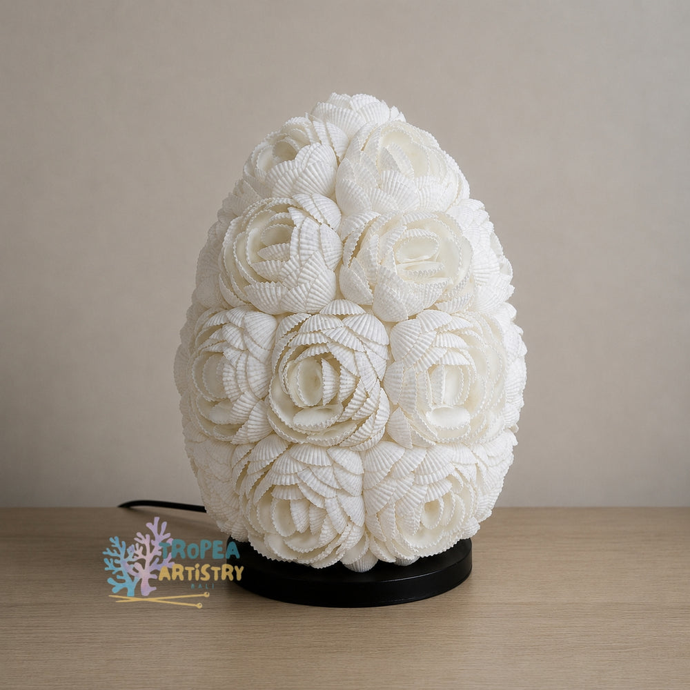 An egg-shaped white fabric sculpture featuring intricate floral and rose patterns, displayed on a black base on a light wood surface against a neutral beige background.