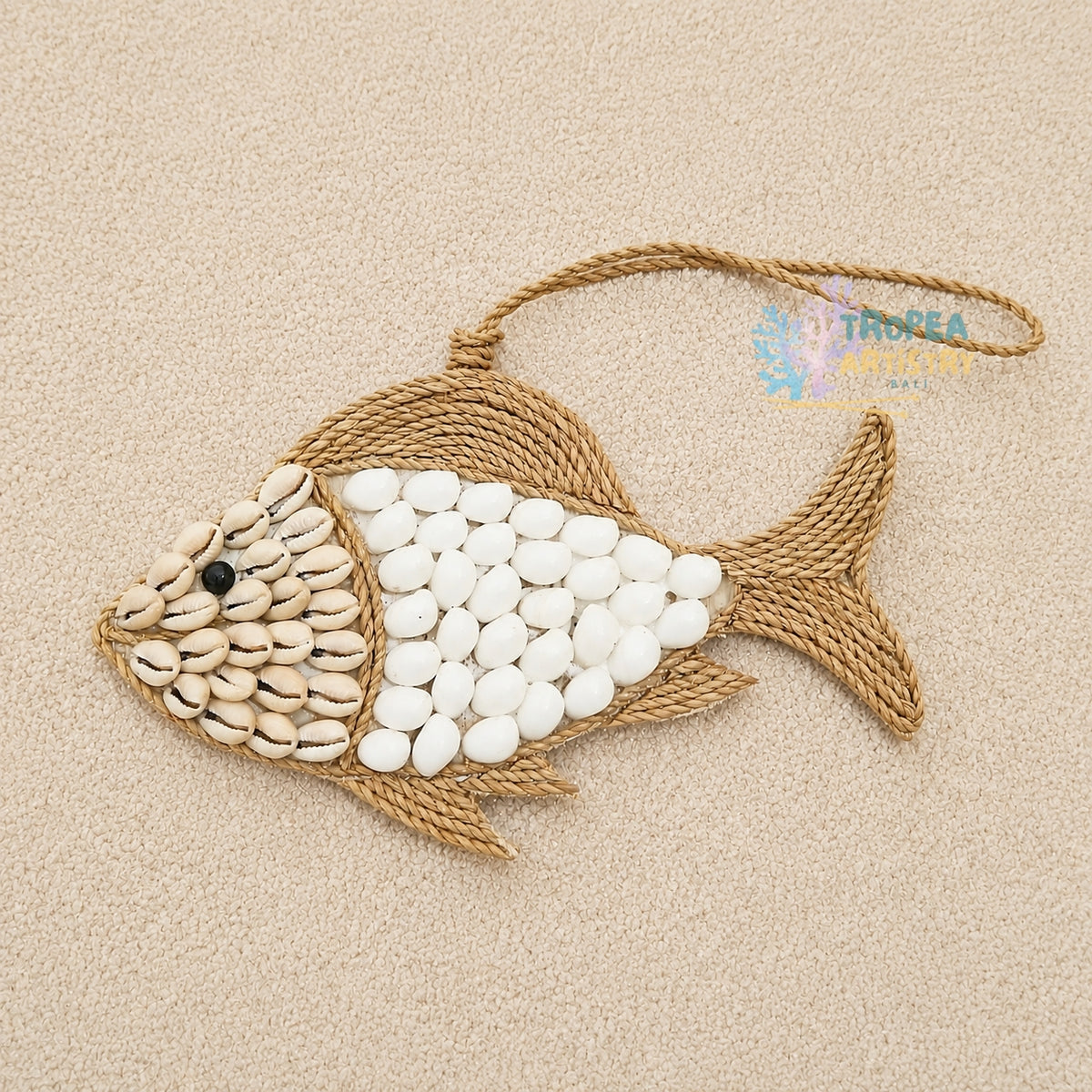 A decorative fish-shaped wall hanging or ornament made from natural rope and white and beige shells, photographed on a light beige textured background. A small black bead eye and a hanging loop are visible.
