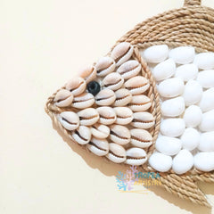 A decorative fish wall art piece made from natural cowrie shells and white pebbles, bordered by woven rope, photographed on a plain light beige background.