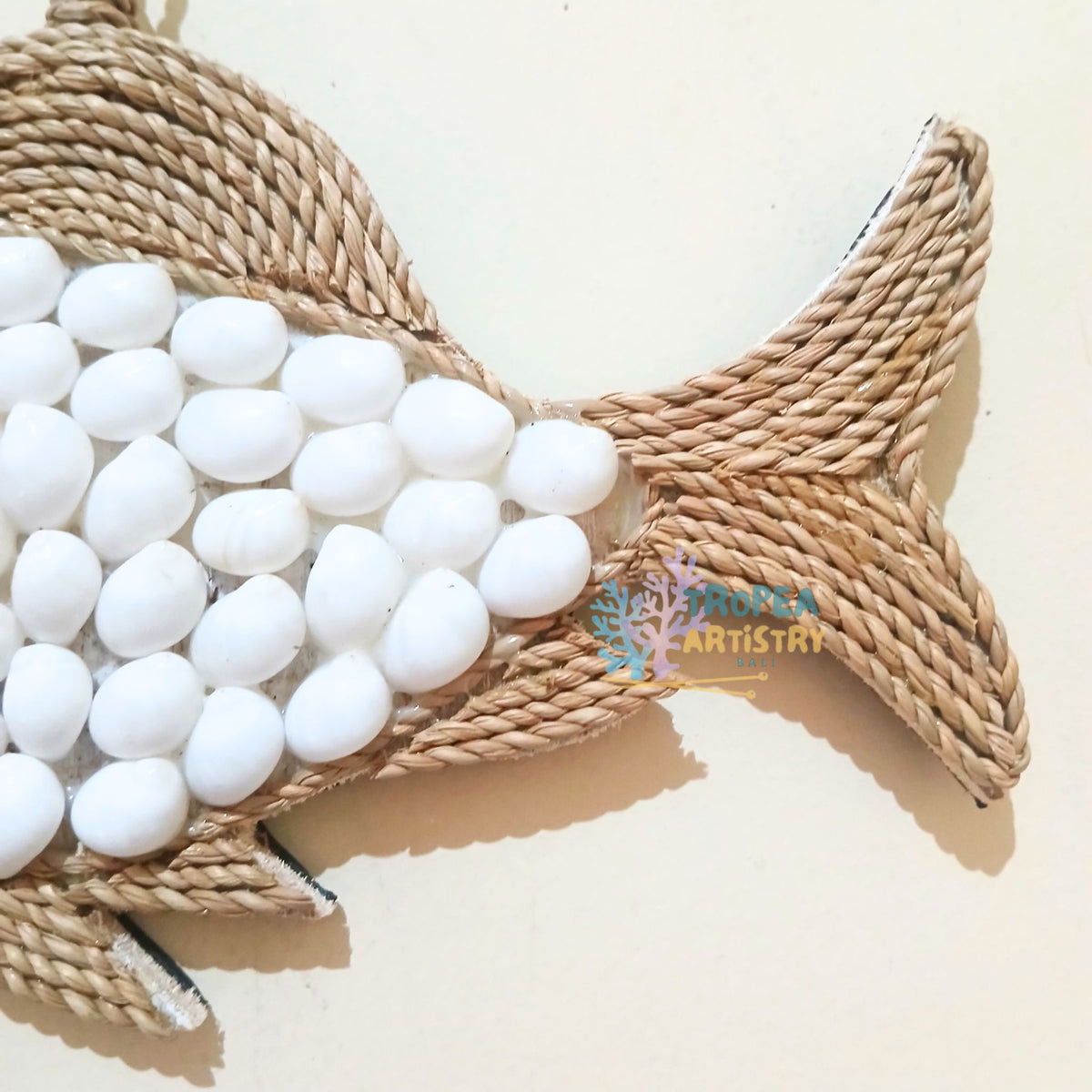 A close-up of a fish-shaped wall hanging or decorative piece made from natural woven rope, with white pebble-like accents along the body, shown against a plain light beige background.