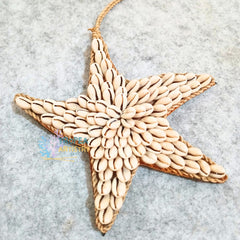 A handmade starfish-shaped wall hanging or ornament made from woven natural fiber and beige cowrie shells, photographed on a light gray textured surface.