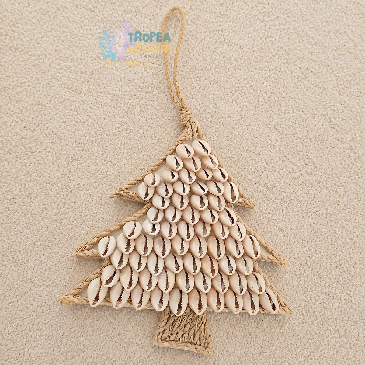 A handmade Christmas tree ornament made from natural jute rope and beige cowrie shells, displayed on a textured beige fabric background.