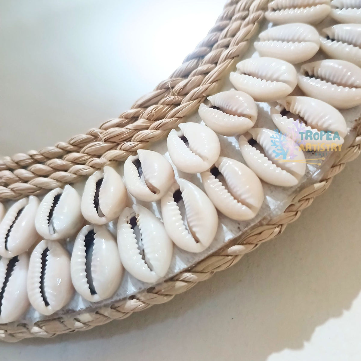 A close-up of a woven natural fiber tray or basket trim decorated with small white cowrie shells arranged in a curved row. The shells have dark slit openings, and the woven base is tan and beige.