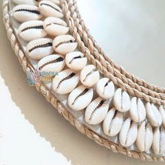 A close-up of a woven natural fiber basket or tray decorated with rows of white cowrie shells, showing the texture of the braided trim and the glossy shell details.