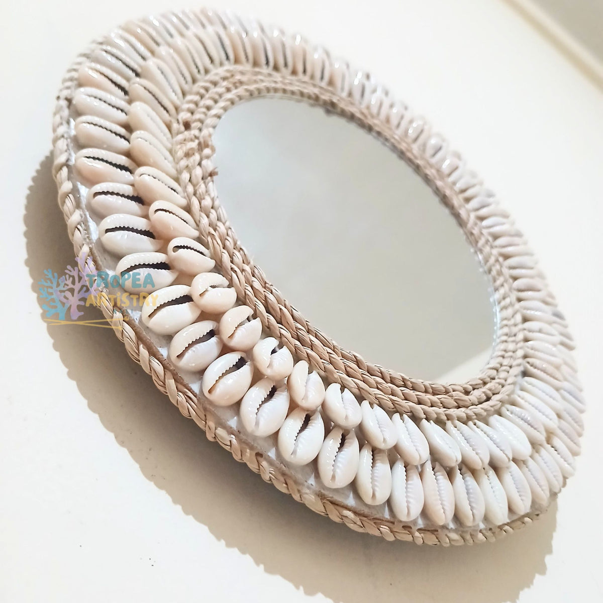 Oval wall mirror with a decorative frame made of woven natural fiber and rows of white cowrie shells, photographed on a light beige surface.