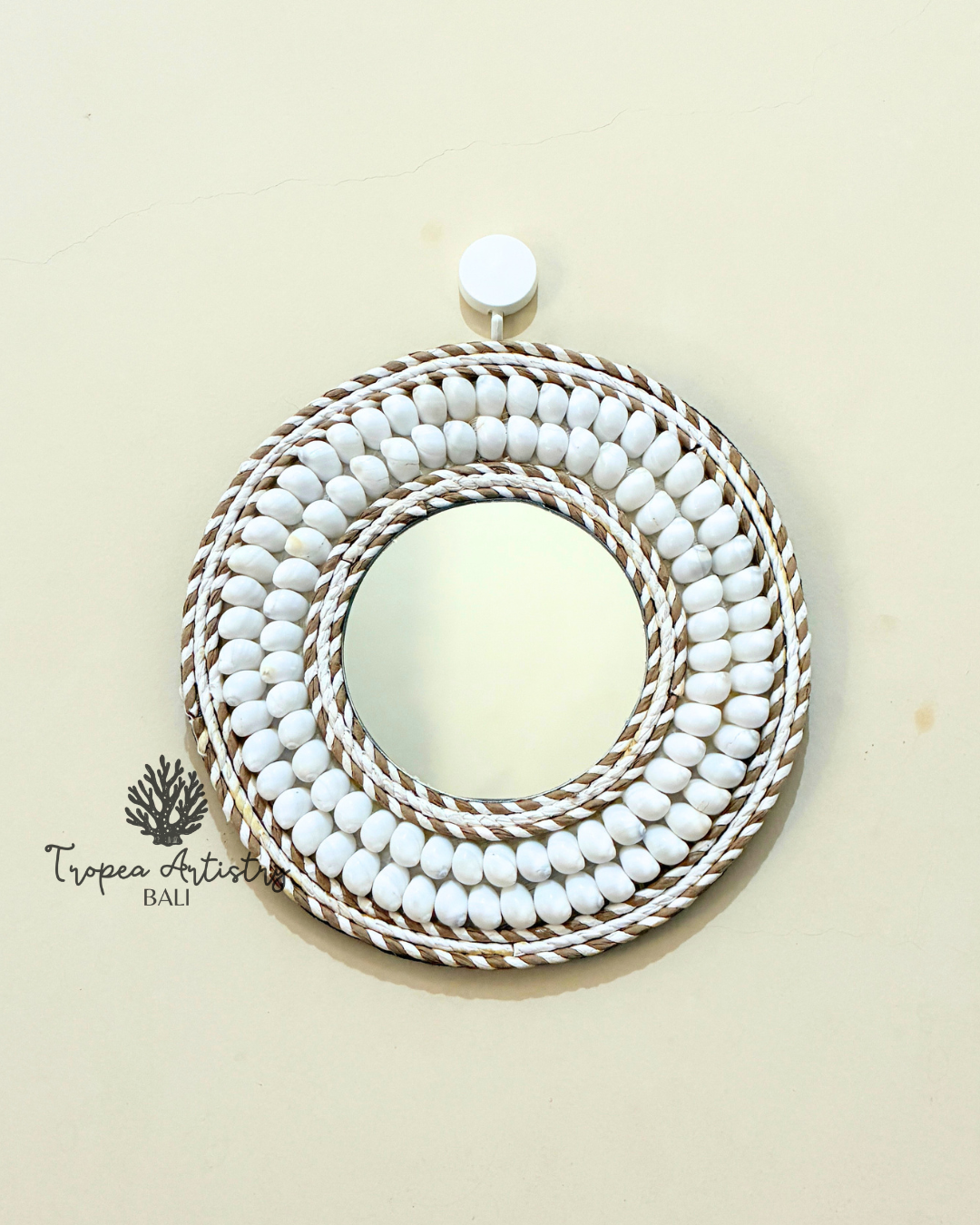 A round decorative mirror with a thick, multi-layered border of white beads and brown-and-white braided rope, hanging on a light beige wall.
