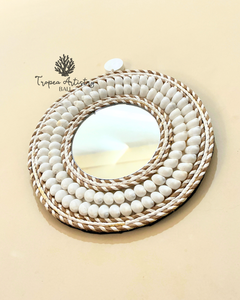 A round decorative mirror with a layered frame made of white beads and braided natural fiber trim, photographed on a warm beige background. A Tropéa Artistry BALL logo is visible in the upper left.