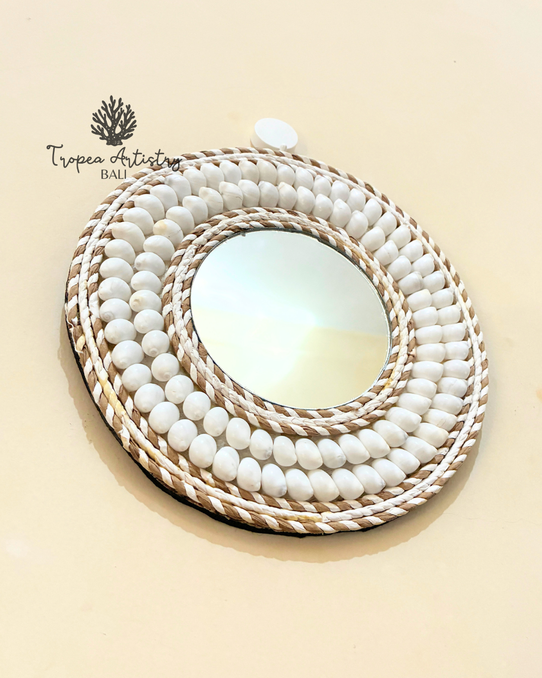An oval wall mirror with a thick, layered circular frame made of white beads and natural woven raffia rope, shown against a solid beige background. A brand logo for 'Tropea Artisan Bali' is visible in the upper left corner.