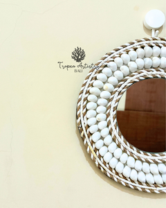 A round decorative mirror with a white beaded frame and braided rope detailing, mounted on a light beige wall. The brand name 'Tropéa Artistry Bali' is printed on the wall to the left.