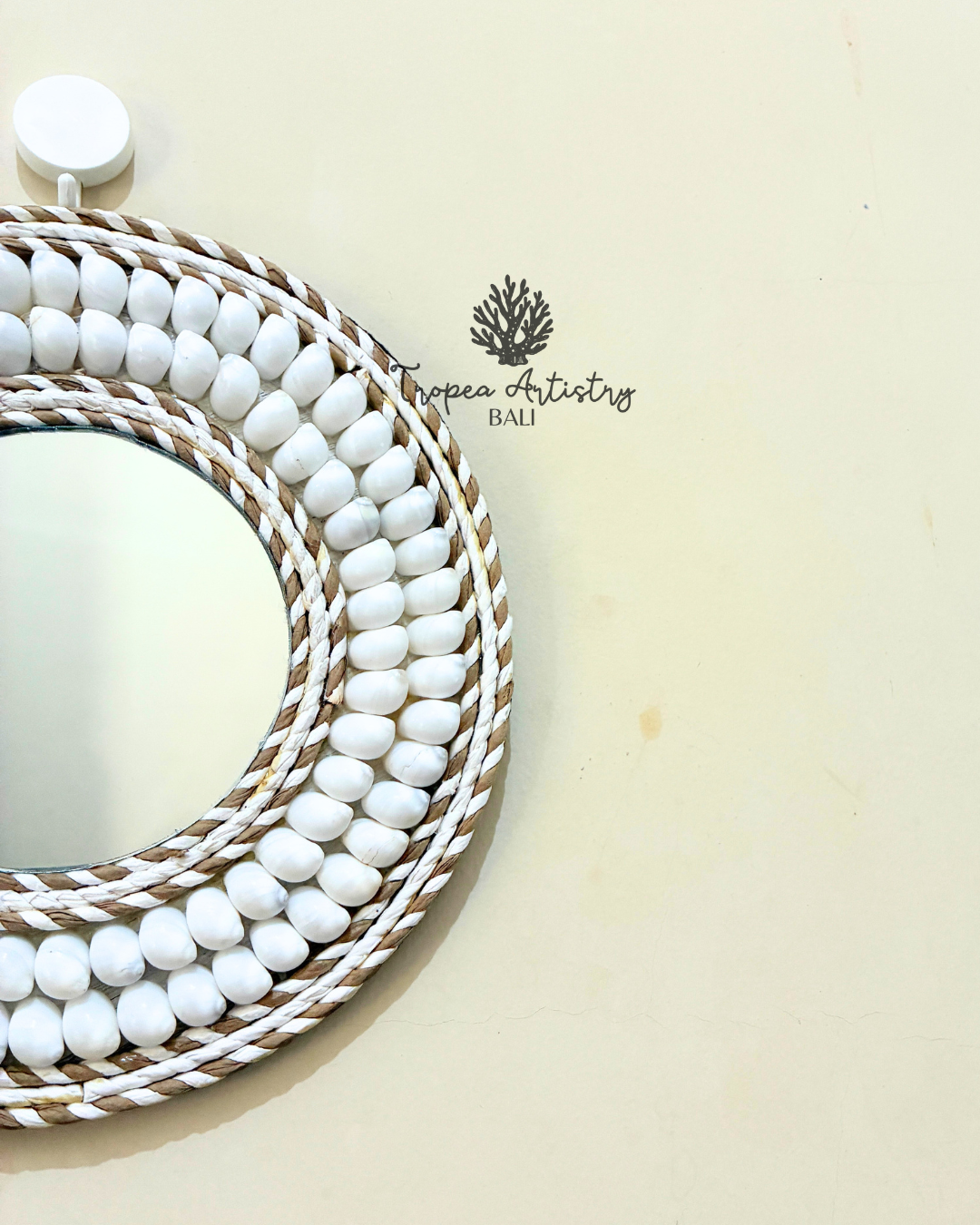 A decorative oval wall mirror with a white beaded frame and braided rope borders, photographed on a warm cream background. A small 'Tropen Artistry Bali' logo is printed on the surface.