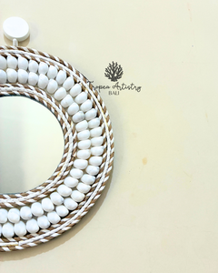 A decorative oval wall mirror with a white beaded frame and braided rope borders, photographed on a warm cream background. A small 'Tropen Artistry Bali' logo is printed on the surface.