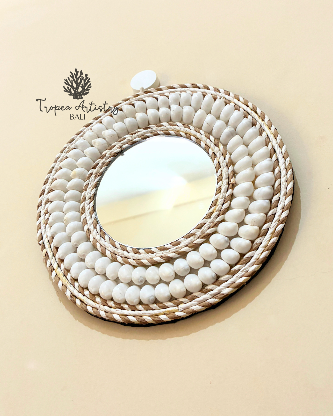 An oval wall mirror with a thick, layered circular frame made of white ceramic beads and natural woven rope, mounted on a beige wall. The brand logo 'Tropéa Artistry Bali' is visible in the upper left corner.