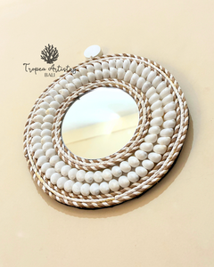An oval wall mirror with a thick, layered circular frame made of white ceramic beads and natural woven rope, mounted on a beige wall. The brand logo 'Tropéa Artistry Bali' is visible in the upper left corner.