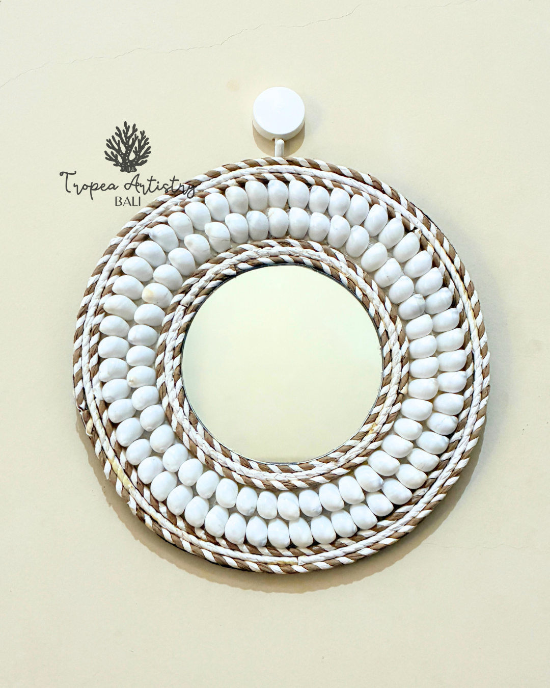A round wall mirror with a decorative frame made of concentric rings of white beads and twisted natural-fiber rope, hanging on a light beige wall. The Tropea Artistry Bali brand logo is visible in the upper left.