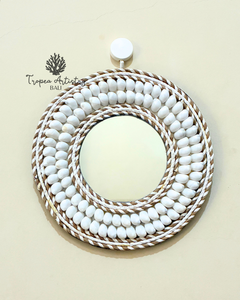 A round wall mirror with a decorative frame made of concentric rings of white beads and twisted natural-fiber rope, hanging on a light beige wall. The Tropea Artistry Bali brand logo is visible in the upper left.