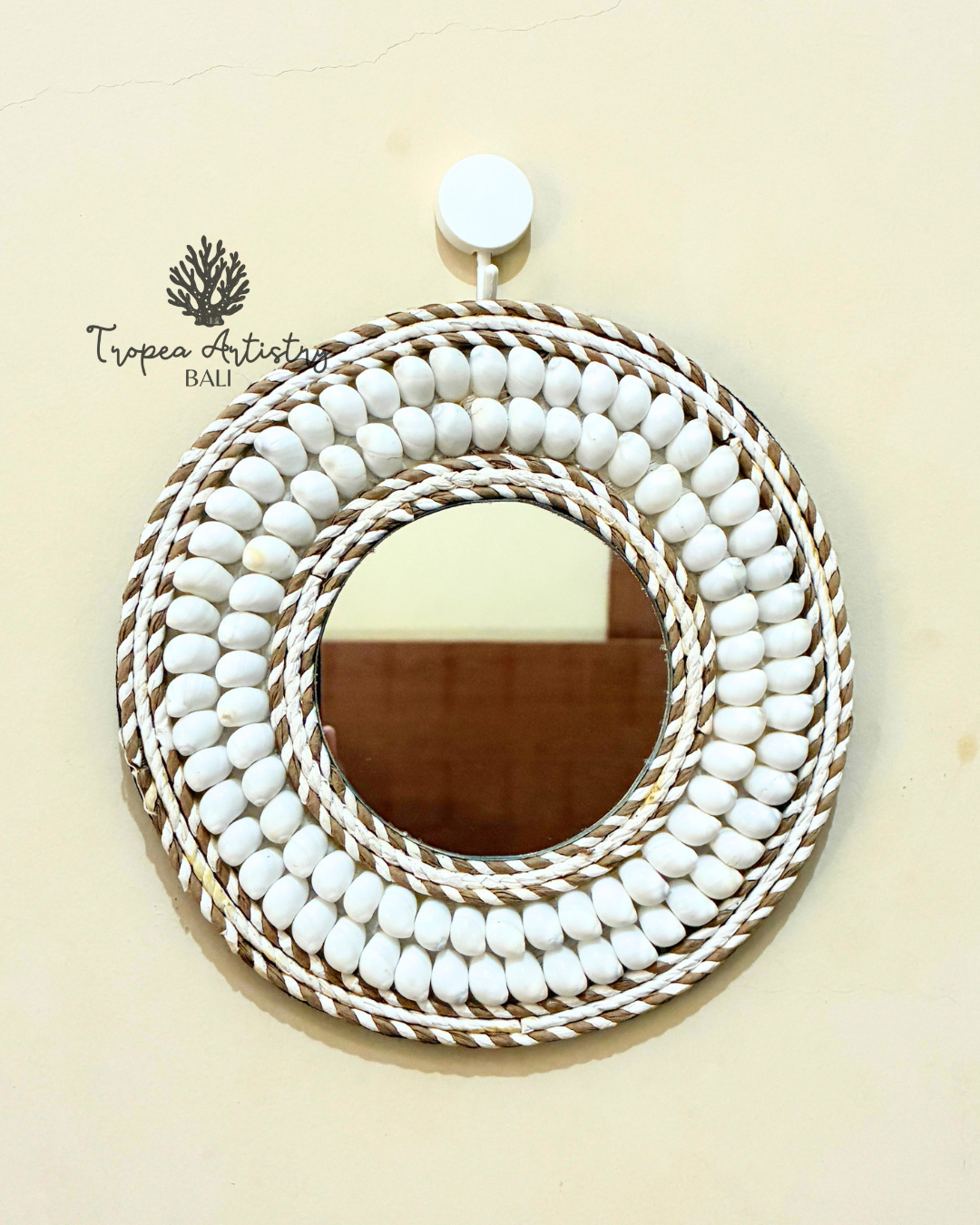 A round decorative mirror with a thick, layered frame made of white painted oval beads and natural tan rope, hanging on a light beige wall. The brand name 'Tropea Artistry Bali' is visible as a watermark on the upper left.