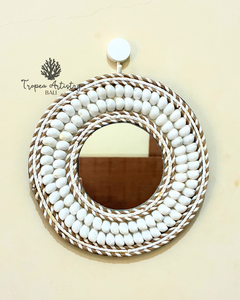A round decorative mirror with a thick, layered frame made of white painted oval beads and natural tan rope, hanging on a light beige wall. The brand name 'Tropea Artistry Bali' is visible as a watermark on the upper left.