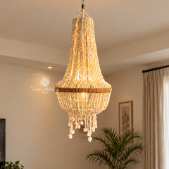 A large, elegant chandelier with cascading crystal beads and a gold-toned band, hanging in a warm, modern interior room with a visible Tropéa Artistry Bali watermark.