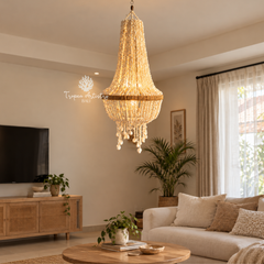 A large, elegant crystal chandelier with a warm gold-toned frame hangs in a beautifully styled living room with a beige sofa, wooden furniture, and potted plants.