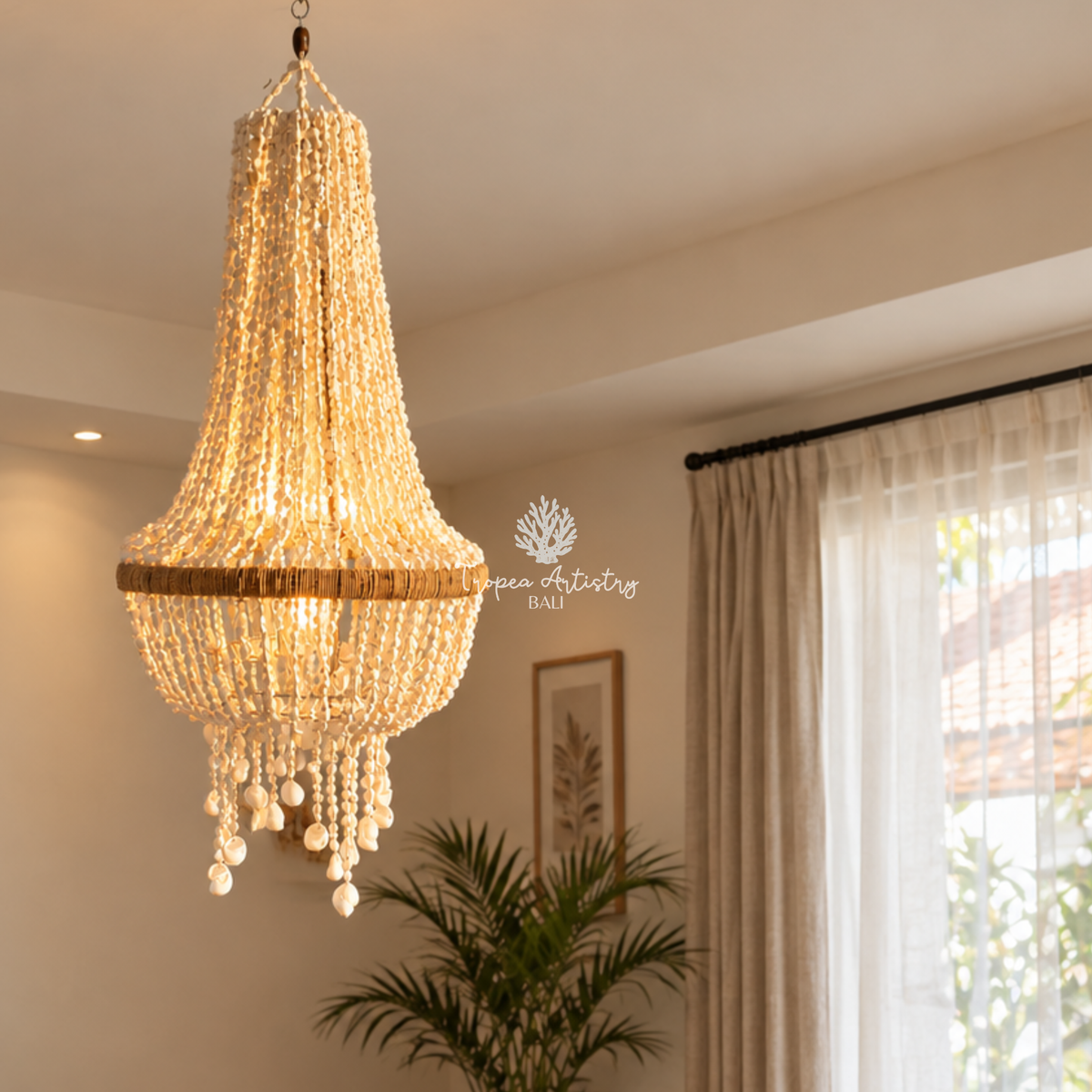 A large, elegant tiered chandelier made of warm white crystal beads and a natural woven rattan band, hanging in a bright, modern room with beige walls, sheer curtains, and a potted plant.