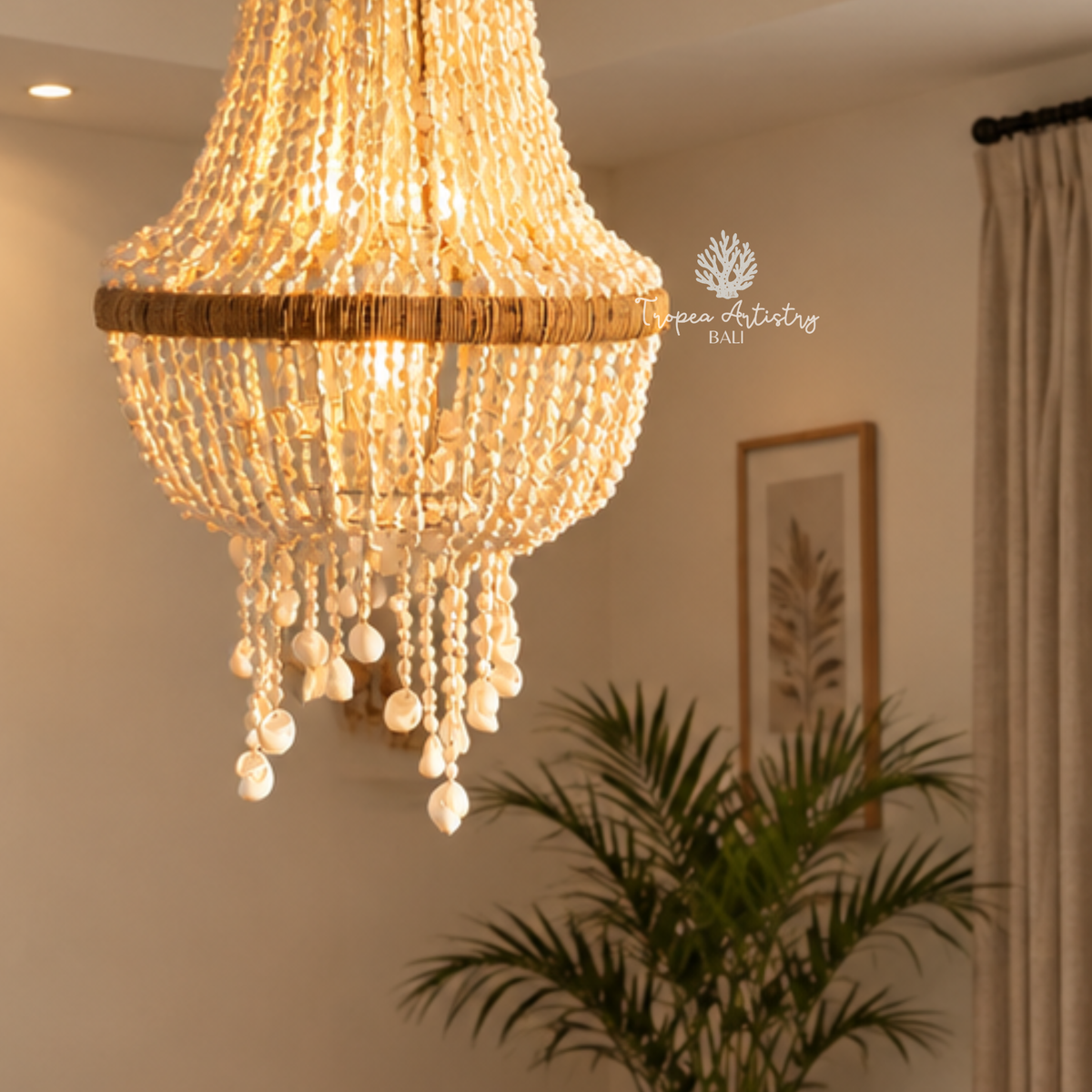A warm, glowing crystal chandelier with hanging pearl-like beads and a natural woven band, shown suspended in a softly lit room with a plant and framed wall art in the background.
