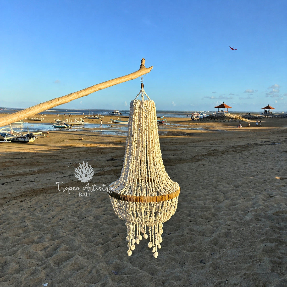 A large decorative chandelier made of white beads and gold-toned metal rings hangs from a wooden branch on a sandy beach, with boats, water, and blue sky in the background.