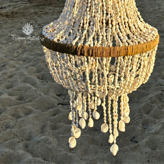 A bohemian-style chandelier made of natural wood and white seashells, hanging on a sandy beach. The brand name 'Tropica Designing Bali' is visible as a watermark in the upper left corner.