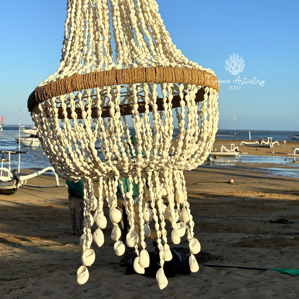 A large decorative chandelier made of woven natural fibers and hanging cowrie shells, photographed outdoors on a sandy beach with boats and water in the background. A watermark reading 'Ocean Artistry Bali' is visible on the right.