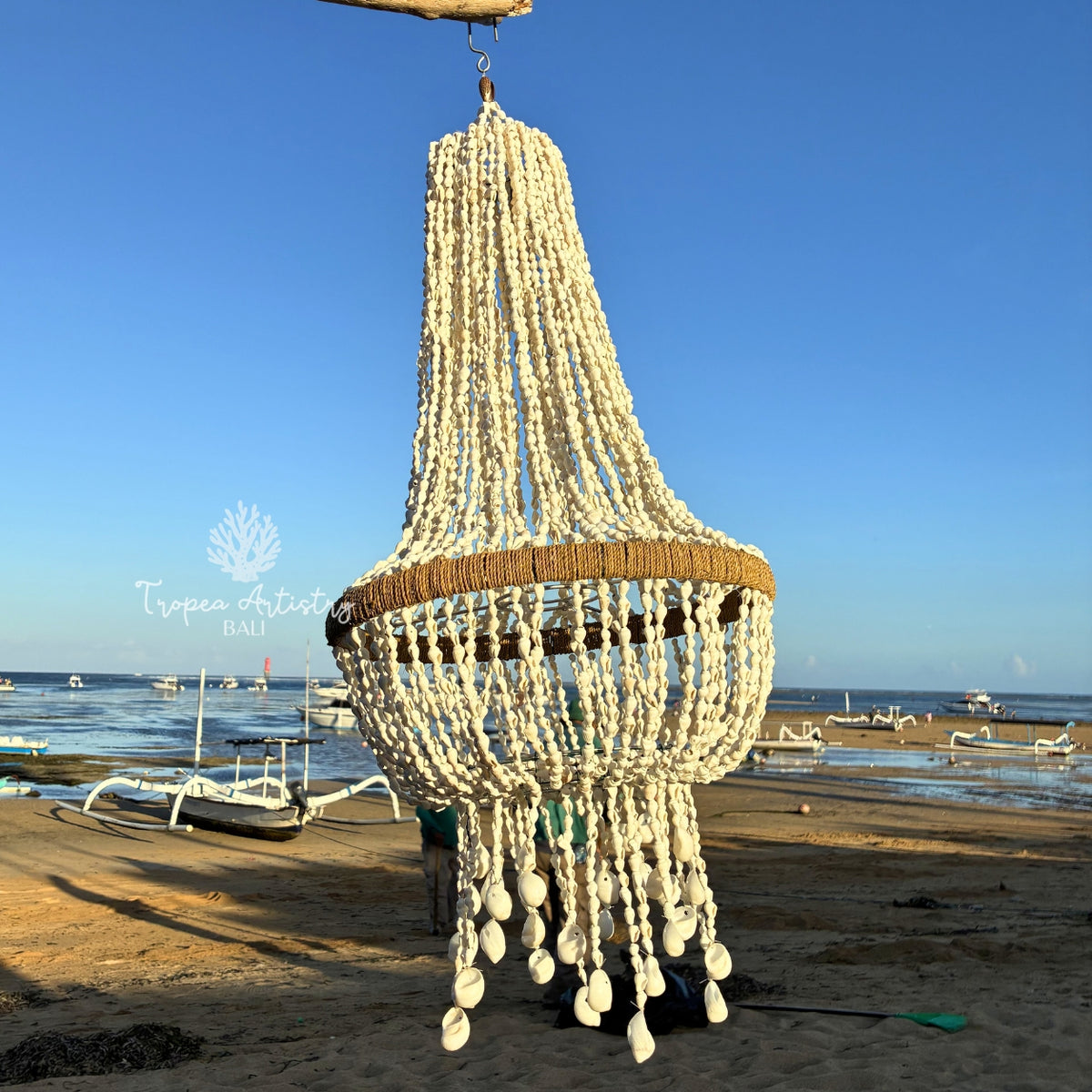 A large bohemian-style chandelier made of off-white shell beads and a natural brown woven ring, hanging outdoors on a beach with a blue sky and boats in the background.
