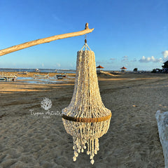A large, tiered beach chandelier made of natural beige seashells and beads hangs from a wooden branch on a sandy beach under a clear blue sky. A watermark for 'Tropen Artistry Bali' is visible on the left.