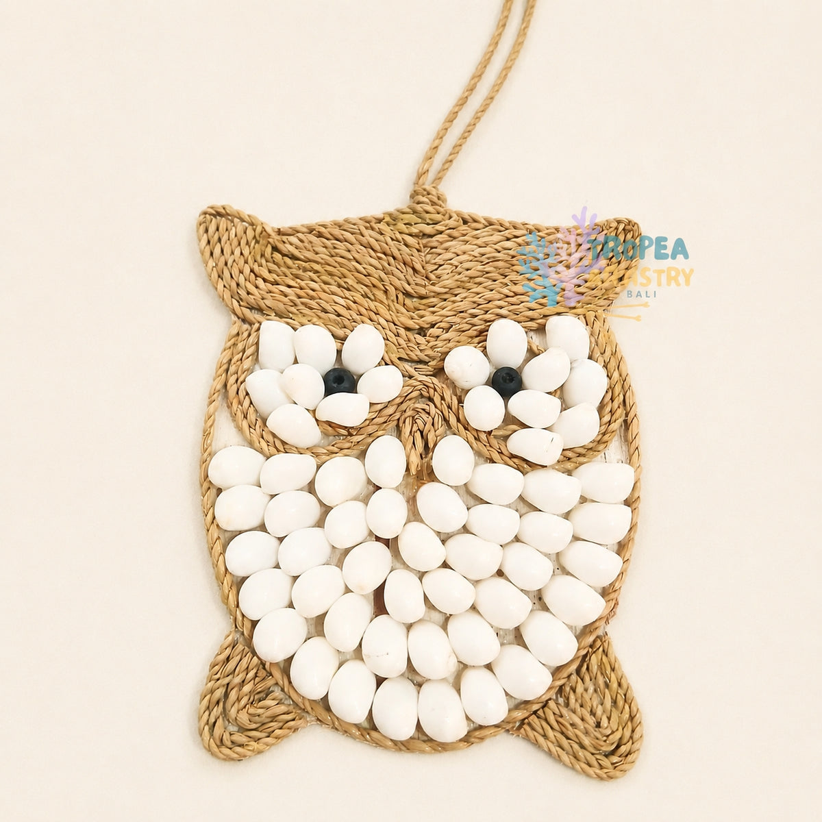 A handmade woven jute owl wall hanging ornament decorated with white seed beads for the eyes and body, featuring black bead eyes and a hanging loop at the top, shown on a light beige background.