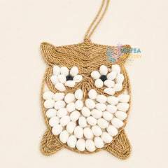 A handmade woven jute owl wall hanging ornament decorated with white seed beads for the eyes and body, featuring black bead eyes and a hanging loop at the top, shown on a light beige background.