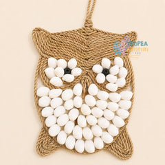 A handmade woven rope owl wall hanging with white pebble eyes and white pebble flower details, shown against a solid light beige background. A 'Tropaea Artistry Bali' logo is visible on the right side.
