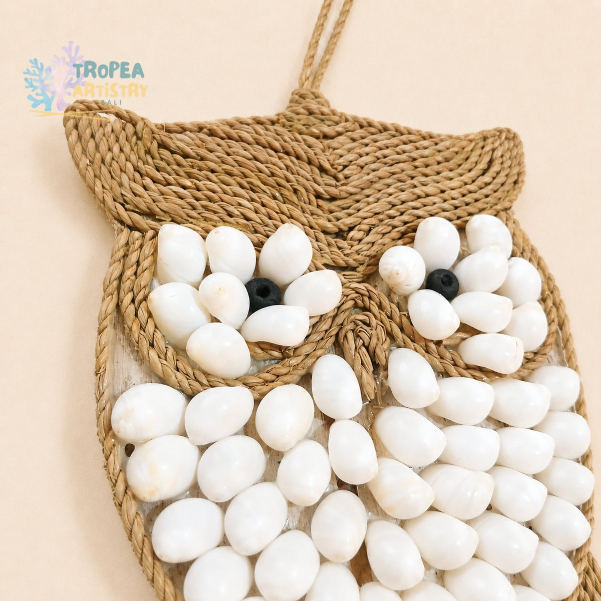 Owl-shaped woven wall hanging made from natural jute rope, decorated with white seashells for the body and eyes, and small black bead eyes, shown against a light beige background.
